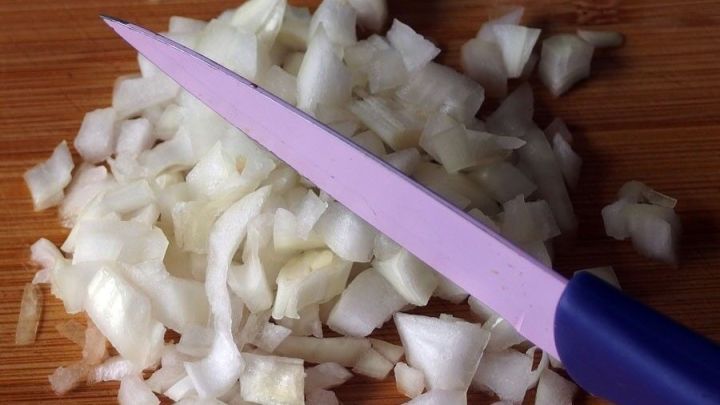 Gehackte Zwiebeln liegen auf einem Holzbrett, w&auml;hrend ein lila Messer daneben zum Schneiden bereitsteht.