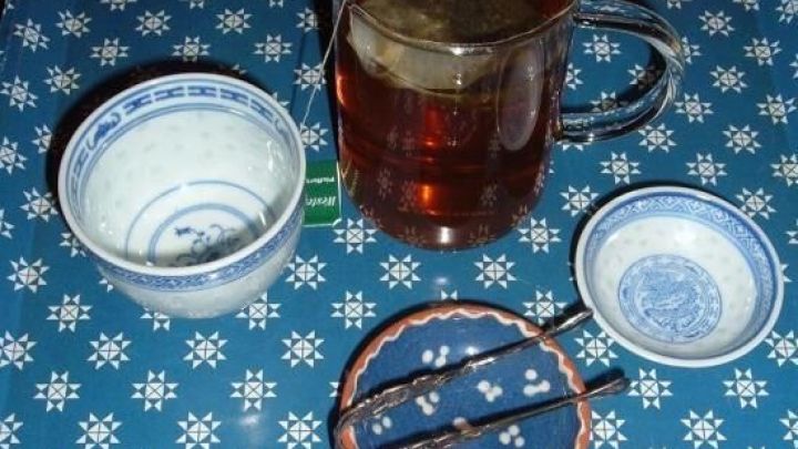 Eine Teetasse mit frisch gebr&uuml;htem Tee steht auf einem blauen Tisch. Nebenan ein kleiner Becher und ein Teesieb.