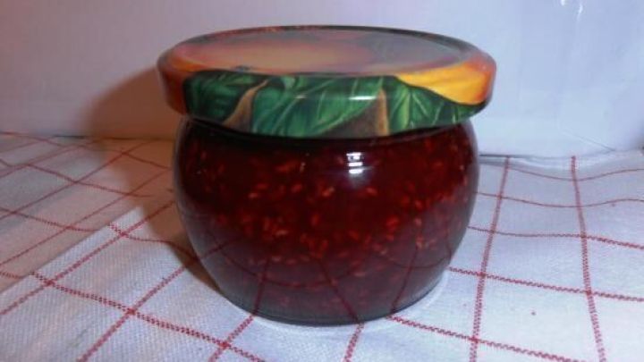 Ein Glas Himbeermarmelade steht auf einem karierten Tuch. Der Deckel hat ein frisches Gr&uuml;n und ansprechendes Design.