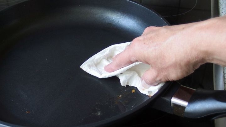 Eine Hand wischt mit einem Tuch &uuml;ber eine Pfanne, um sie nach dem R&uuml;hrei reinigen. Die Pfanne liegt auf einem Herd.