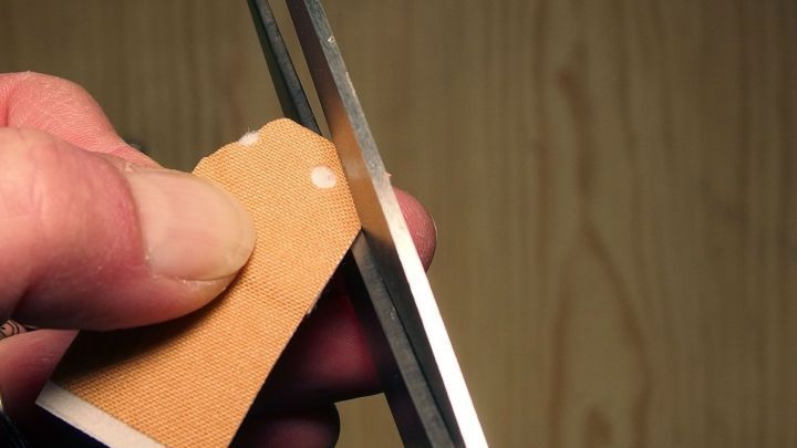 Eine Hand h&auml;lt ein Pflaster und schneidet es sorgf&auml;ltig mit einer Schere, w&auml;hrend eine Holzoberfl&auml;che sichtbar ist.