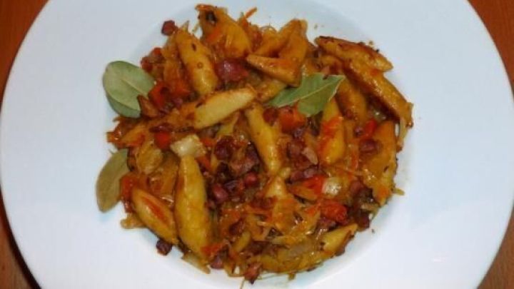 Knusprig gebratene Schupfnudeln liegen goldbraun auf einem wei&szlig;en Teller, garniert mit gr&uuml;nen Bl&auml;ttern und etwas Speck.