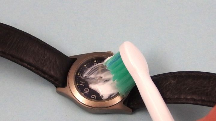 Eine Uhr wird mit einer Zahnb&uuml;rste gereinigt, w&auml;hrend sie auf einem blauen Hintergrund liegt, um Kratzer zu entfernen.