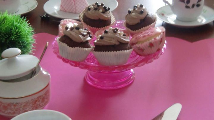 Auf einer rosa Tablett befinden sich schokoladige Muffins mit Sahne und Schokoladenst&uuml;ckchen, umgeben von zierlichen Tassen.
