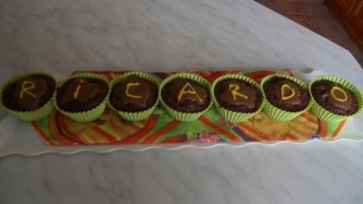 Schokoladenmuffins in gr&uuml;nen F&ouml;rmchen auf Tablett, dekoriert mit gelben Buchstaben "RICARDO" in einer K&uuml;chenumgebung.
