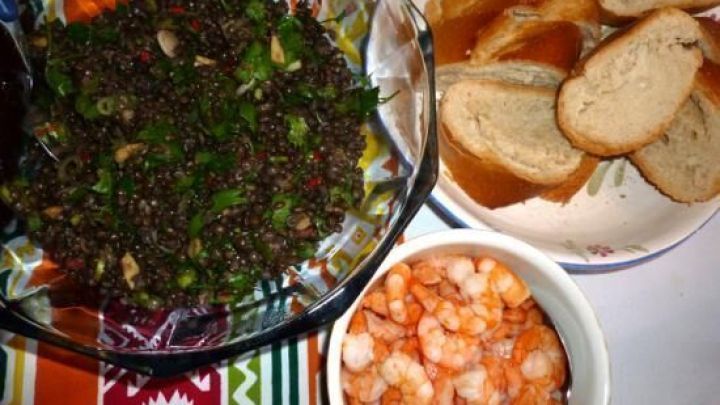 Ein bunten Salat aus Belugalinsen wird in einer Sch&uuml;ssel serviert, neben Shrimps und Brot auf einem gemusterten Tisch.