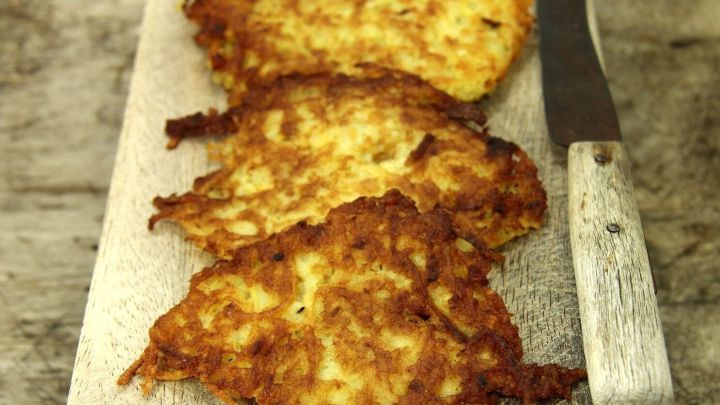Knusprige Kartoffelr&ouml;sti mit Fr&uuml;hlingszwiebeln liegen auf einem rustikalen Holzbrett neben einem K&uuml;chenmesser.
