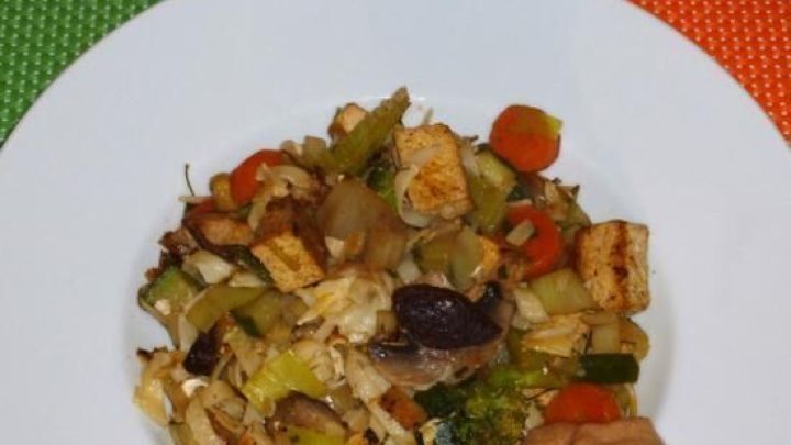 Eine bunte Bami-Pfanne mit viel Gem&uuml;se und einer Erdnusssauce auf einem wei&szlig;en Teller vor einem bunten Hintergrund.