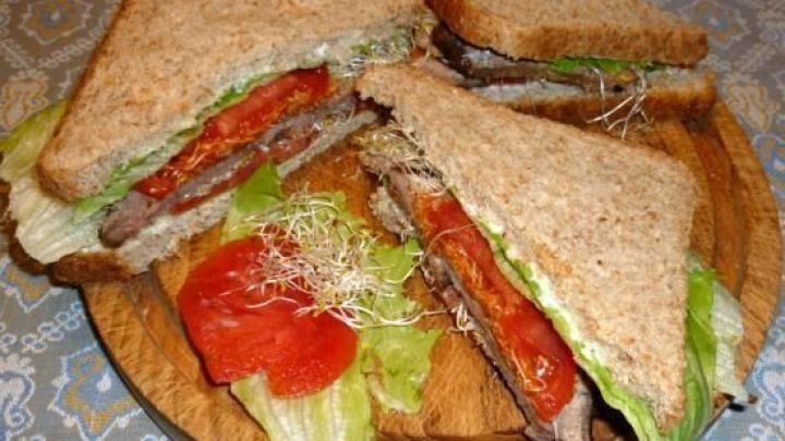 Leckere Sandwiches mit Bratenresten, belegt mit Salat und Tomate, auf einem Holzbrett angerichtet.
