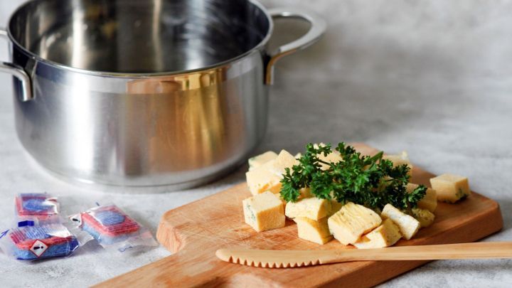 Ein Schneidebrett mit gew&uuml;rfeltem Eierstich und frischer Petersilie liegt neben einem leeren Topf auf einer Fl&auml;che.