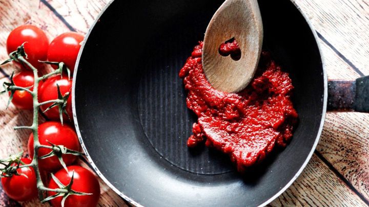 In einer Pfanne wird Tomatenmark mit einem Holzl&ouml;ffel verr&uuml;hrt, w&auml;hrend frische Tomaten am Rand liegen.