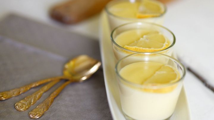 Drei Gl&auml;ser mit Zitronen-Vanille-Creme stehen auf einer wei&szlig;en Platte, umgeben von Besteck auf einem grauen Untergrund.