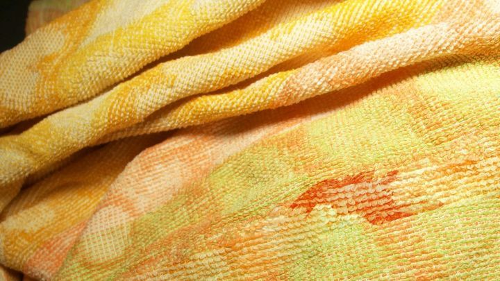 Ein gelbes Tuch liegt gefaltet, sichtbar mit einem Rotweinfleck darauf, in einem dunklen Umfeld.