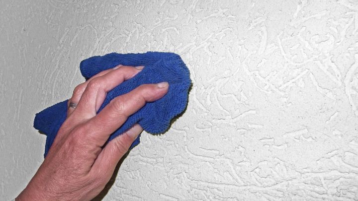 Eine Hand mit einem blauen Microfasertuch wischt M&uuml;ckenflecken von einer strukturierten wei&szlig;en Wand ab.