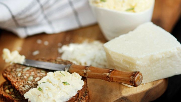 K&auml;sebutter wird auf dunklem Brot mit einem Messer verteilt, w&auml;hrend ein St&uuml;ck Parmesan daneben liegt.