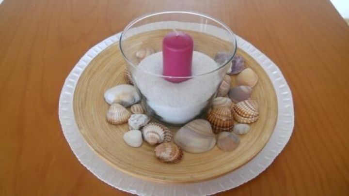 Eine pinke Kerze steht in einem Glas auf Sand, umgeben von dekorativen Muscheln auf einem Holztablett.