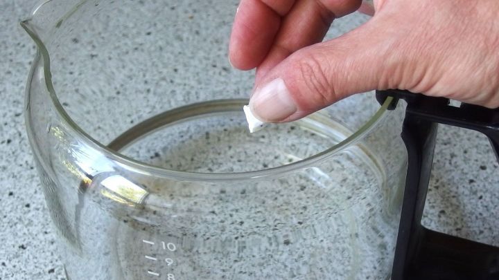 Eine Hand legt ein St&uuml;ck Reinigungsmittel in eine eingebrannte Glaskanne, w&auml;hrend sie auf einer grauen Arbeitsplatte steht.