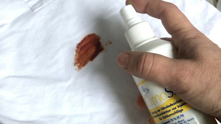 Ein Hand Spray entfernt einen roten Marinadefleck von einem wei&szlig;en Kleidungsst&uuml;ck auf einem Holzuntergrund.
