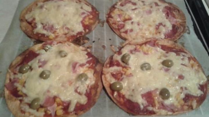 Vier Mini-Pizzas mit K&auml;se, Oliven und Schinken backen auf einem Blech im Ofen, auf Backpapier liegend.
