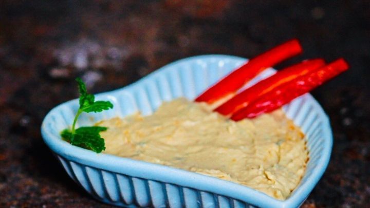 Ein Herzsch&auml;lchen mit Hummus Dip wird pr&auml;sentiert, garniert mit frischen Paprikastreifen und einem Blatt Koriander.