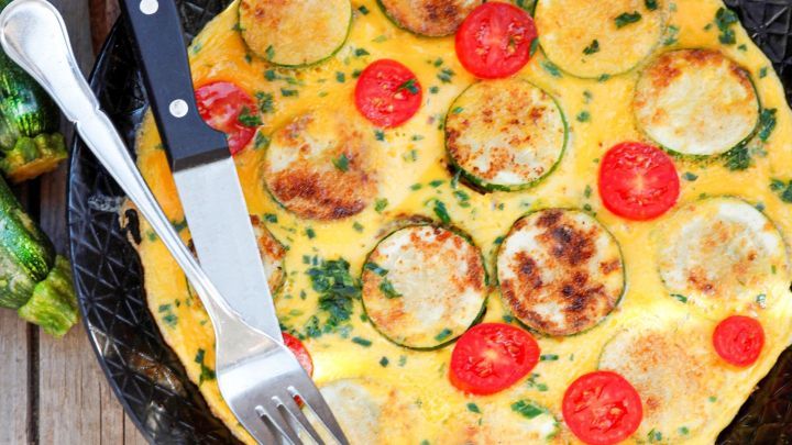 Ein Omelette, belegt mit Zucchini und Tomaten, liegt auf einem schwarzen Teller, w&auml;hrend Besteck daneben bereitliegt.