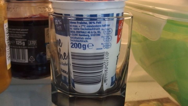 Ein Sahnebecher steht stabil in einem Glas im K&uuml;hlschrank, um ein Umkippen zu verhindern. Im Hintergrund sind Lebensmittel zu sehen.