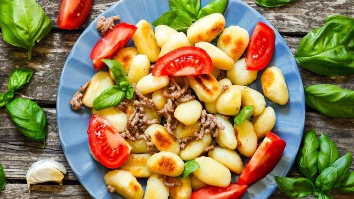 Eine bunte Gnocchi-Pfanne mit Hackfleisch und frischen Tomaten liegt auf einem blauen Teller, umgeben von Basilikumbl&auml;ttern.