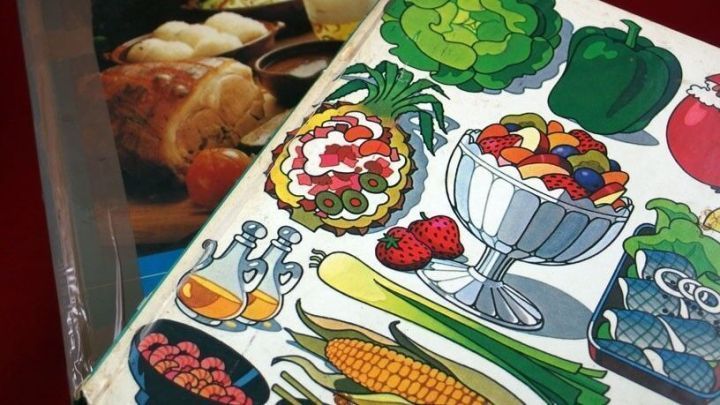 Ein Kochbuch mit bunten Illustrationen von Lebensmitteln liegt auf einem Tisch, umgeben von anderen Kochb&uuml;chern und Gerichten.