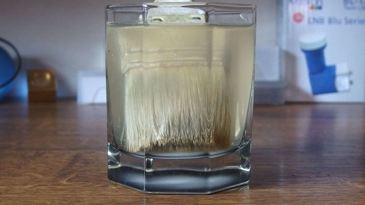 Ein Pinsel wird in ein Glas mit L&ouml;sungsmittel getaucht, um eingetrocknete Acrylfarbe zu entfernen. Der Tisch ist aus Holz.