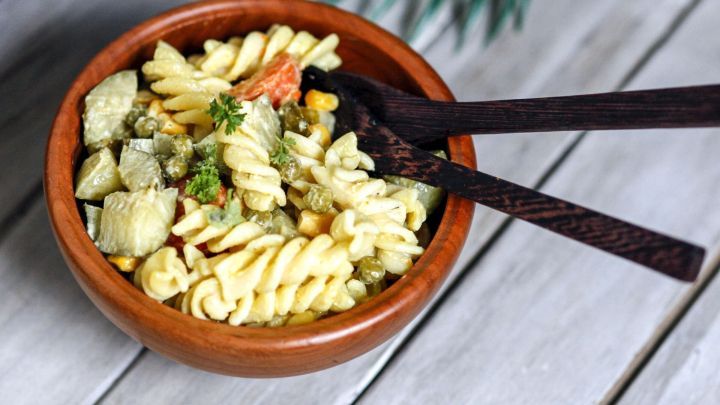 Ein bunter Nudelsalat in einer Holzsch&uuml;ssel wird mit zwei Holzl&ouml;ffeln serviert, umgeben von einem rustikalen Holzuntergrund.
