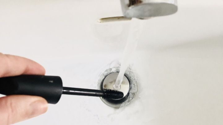 Ein Hand ist mit einer Wimperntusche &uuml;ber einem Waschbecken, w&auml;hrend Wasser aus dem Wasserhahn flie&szlig;t.