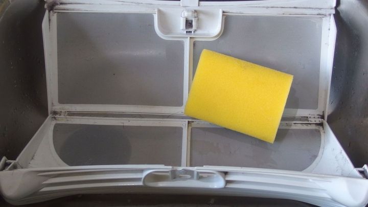 Ein W&auml;schetrockner-Fusselsieb liegt ge&ouml;ffnet auf einem Tisch, bereit zur Reinigung mit einem gelben Schwamm.