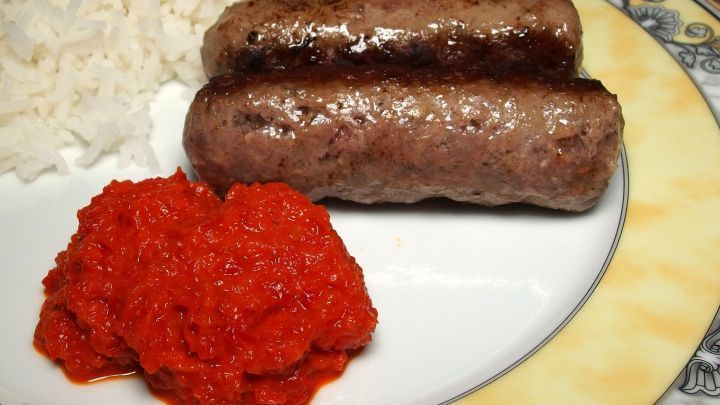 Grillw&uuml;rste liegen auf einem Teller neben Ajvar und Reis, serviert in einer gem&uuml;tlichen Essumgebung.