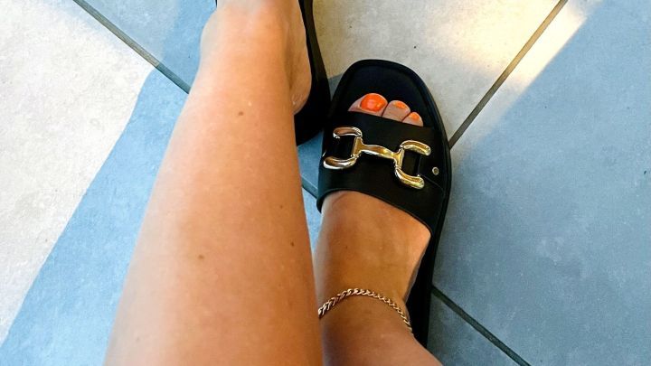 Elegante Beine tragen schwarze Schuhe mit goldenen Details, die auf hellen Fliesen stehen und Licht reflektieren.