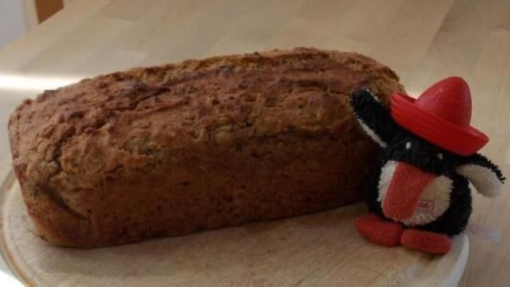 Ein frisch gebackenes Haferflocken-Honig-Brot liegt neben einem lustigen Stofftier mit einem roten Hut auf einem Holzbrett.