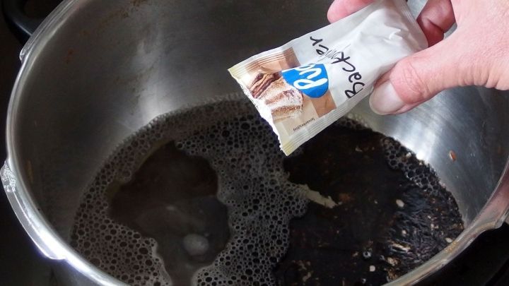 Ein Hand h&auml;lt ein P&auml;ckchen &uuml;ber einen Topf mit verbranntem Inhalt, um ihn zu reinigen.