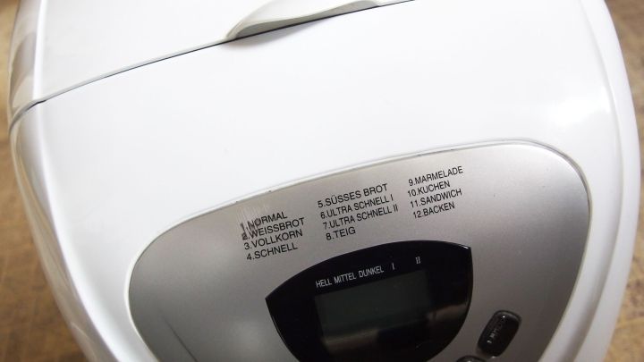 Eine wei&szlig;e Brotbackmaschine steht auf einer Arbeitsfl&auml;che und zeigt Auswahlm&ouml;glichkeiten sowie ein digitales Display f&uuml;r die Programmsteuerung.