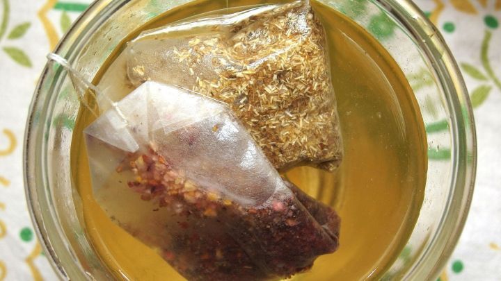 Zwei Teebeutel schwimmen in einem Glas mit goldenem Eistee, auf einem bunten, gemusterten Tisch.