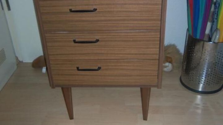 Ein holzfarbener Kommode mit vier Schubladen steht in einem hellen Raum neben einem St&auml;nder mit Regenschirmen.