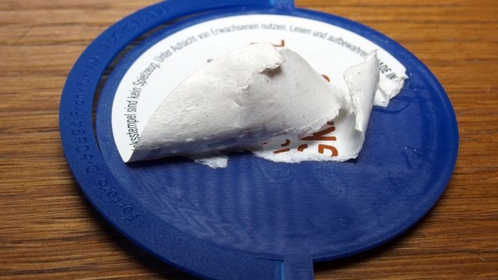 Ein blauer Deckel mit einem St&uuml;ck einer abgel&ouml;sten Etikette, umgeben von einer Holzoberfl&auml;che, zeigt den Prozess der Etikettenentfernung.