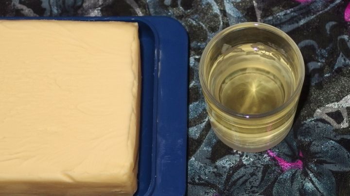 Ein St&uuml;ck Butter liegt auf einem blauen Teller neben einem Glas &Ouml;l, umgeben von einem farbenfrohen, floralem Tischmuster.