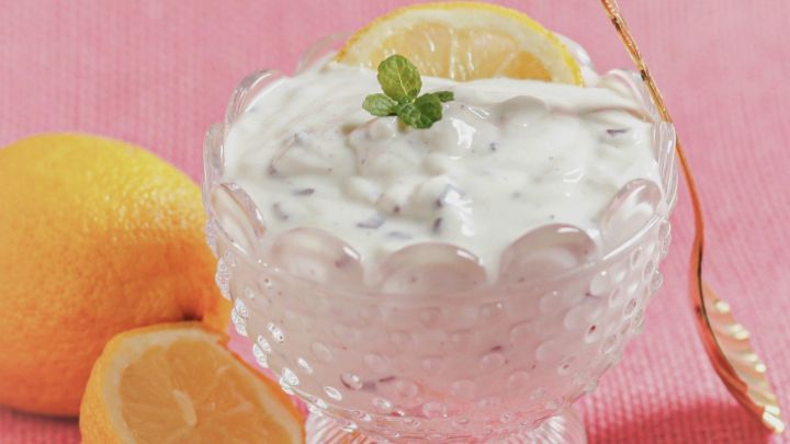 Eine Schale mit Stracciatella Quarkcreme steht auf einem pinken Tuch, umgeben von frischen Zitronen und einem goldenen L&ouml;ffel.