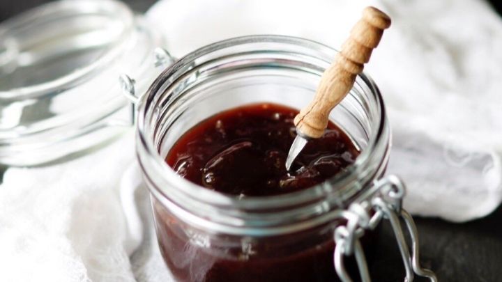 Eine gl&auml;serne Verpackung mit Rosenmarmelade steht auf einem wei&szlig;en Tuch, umgeben von zarten rosa Blumen im Hintergrund.
