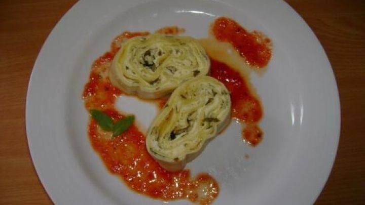 Eine Basilikum-Nudelrolle liegt auf einem wei&szlig;en Teller, umgeben von roter So&szlig;e, die dekorativ angerichtet ist.