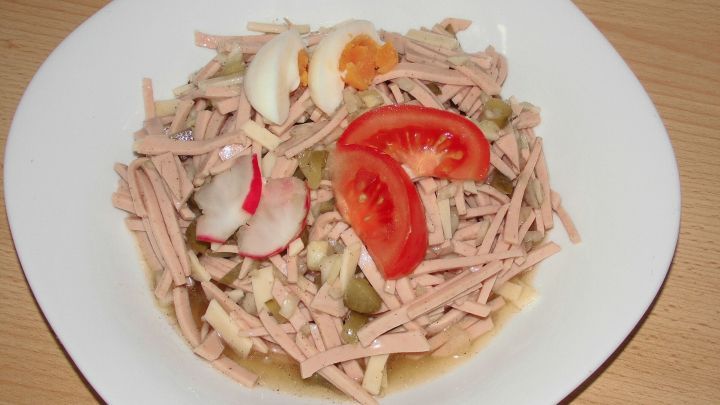 Eine Sch&uuml;ssel Wurstsalat mit Gurkenaufguss, garniert mit Tomatenscheiben, Radieschen und Eiern, auf einem holzfarbenen Tisch.