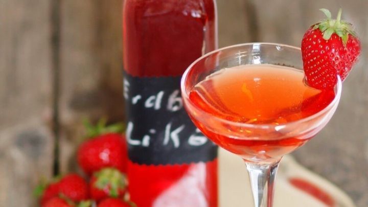 Ein Glas Erdbeerlik&ouml;r steht auf einem silbernen Teller neben frischen Erdbeeren und Zimtstangen auf einem Holzuntergrund.