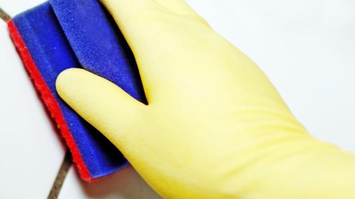 Eine gelbe Gummihandschuhhand putzt mit einem Schwamm Fliesen, w&auml;hrend sie in einem hellen Badfliesenbereich sind.