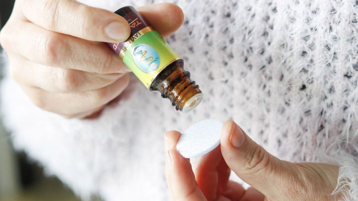 Eine Hand h&auml;lt eine Flasche Lavendel&ouml;l &uuml;ber ein Filzpl&auml;ttchen, um es damit zu befeuchten. Die Umgebung ist hell und l&auml;ssig.