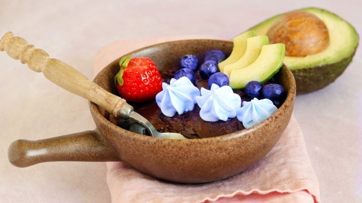 Eine Sch&uuml;ssel mit Avocado-Schoko-Mousse wird dekoriert mit frischen Beeren und einem St&uuml;ck Avocado, auf einem Tuch.