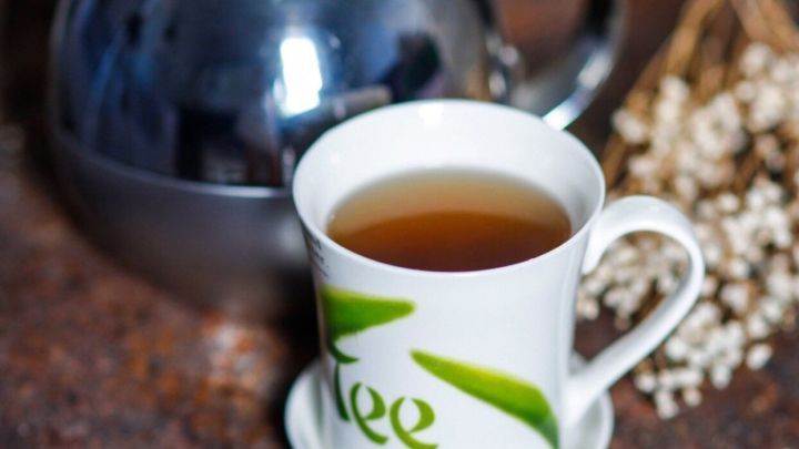 Eine Tasse gr&uuml;ner Tee steht vor einem gl&auml;nzenden Kessel auf einer Holzoberfl&auml;che, umgeben von zarten Pflanzen.